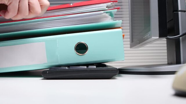 Hand placing a red file on top of a stack of filled binders. Creating an overwhelming workload on a desk next to a computer monitor. Illustrating document management and office organization challenges