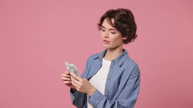 Happy accountant young woman she wears blue shirt white t-shirt casual clothes hold in hand count stack of cash money in dollar banknotes isolated on plain pastel light pink background studio portrait