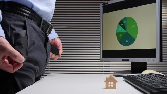 Businessman with empty pockets standing at a desk with a computer displaying a pie chart and a small wooden house, representing financial struggles and uncertain real estate future