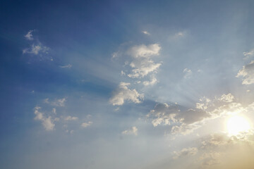 Obraz premium Bright blue sky filled with scattered white clouds and sunlight shining through the clouds. The sun is low enough to emit visible rays in the blue and white sky.