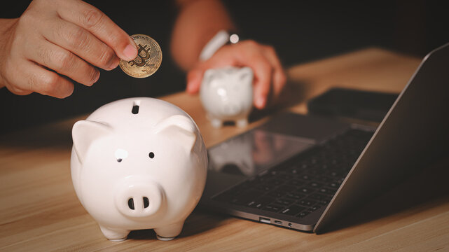 Hand putting physical gold bitcoin into a white piggy bank on a wooden desk with a laptop. Concept of saving digital currency, modern investment, cryptocurrency trading, and future financial wealth.