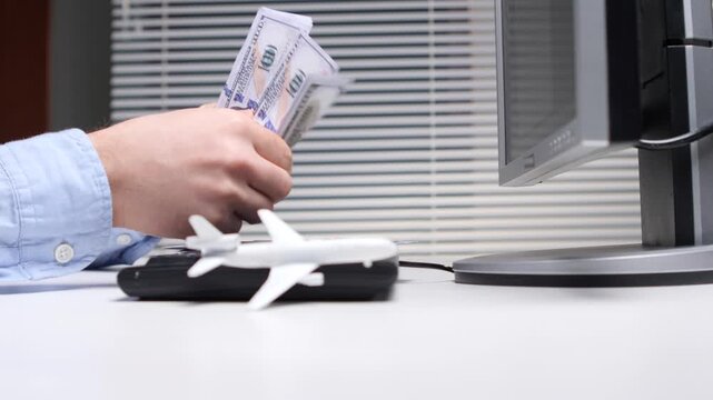 Person's hand holding dollar banknotes while sitting at a desk with a computer monitor and a small airplane model, conceptualizing online travel booking, flight reservations, and vacation expenses