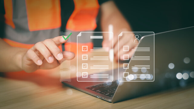 Professional engineer in safety vest using a futuristic digital checklist interface over laptop for quality control, project management, and construction site safety audit process.