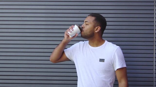 African American man holding paper cup and drinking coffee against wall background, young guy drinking tea and smiling outdoors