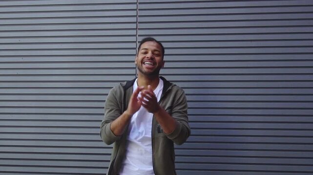 African American man points at the camera and mocks outdoors, guy claps, jokes, and laughs against wall