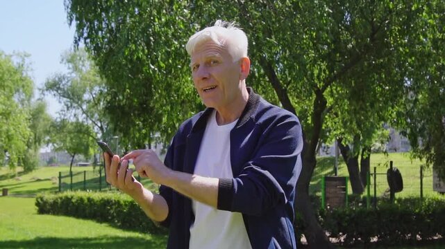 elderly man uses smartphone and gives thumbs-up in the park, gray-haired pensioner came up with idea and solution online outdoors