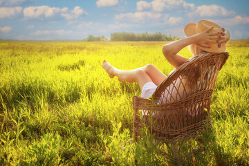 Woman in straw hat relaxing in wicker chair in sunlit grassy field