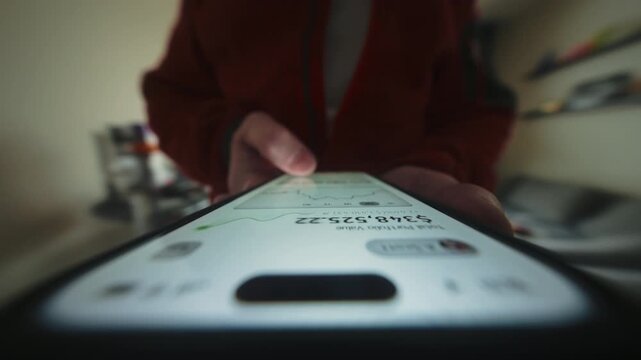 Close-up wide angle male hand holding smartphone. Man walks in apartment using phone. Financial app showing portfolio growth and rising chart. Camera locked at device screen, rotating parallax effect
