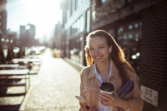 Smiling woman with coffee and smartphone walking on sunny city street