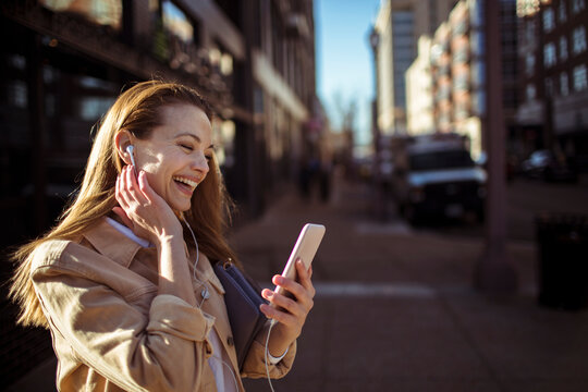 Young woman laughing while listening to music on smartphone in city street