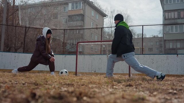 white teens battling for ball at goal, low angle on rough turf, defender lunging and attacker dribbling, gritty outdoor match vibe, apartment blocks and net in frame, high energy and determination