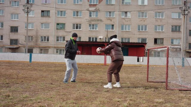 goalkeeper drill with white coach and senior woman, focused saves and crouched stance near net, urban housing fa ade backdrop, overcast sky, padded coats and sneakers, repetition to build reflexes
