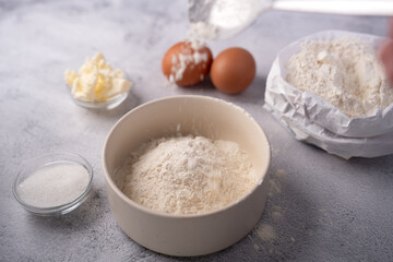 close-up of baking ingredients on a textured tabletop: a ceramic bowl heaped with flour being dusted from a spoon, with eggs, butter, sugar and a crumpled paper bag of flour in soft neutral light