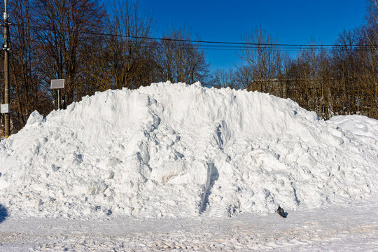 Massive snowdrift, cleared by machinery, dominates the urban street with bare trees and clear blue sky in background