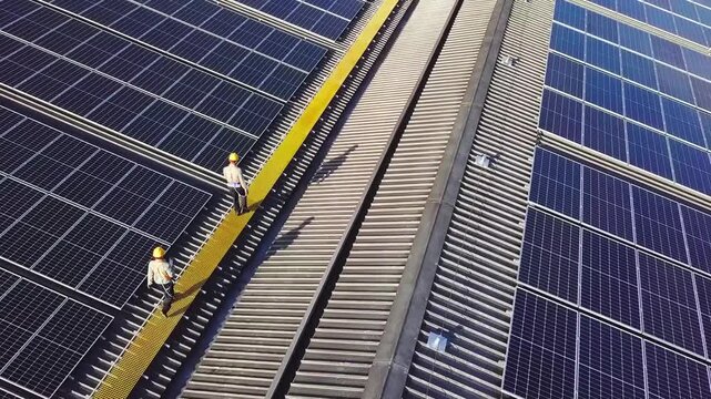 High-angle view of two engineers in yellow hard hats walking on a walkway to inspect a massive solar panel array on a rooftop. Concept of renewable energy, maintenance, and sustainable power.