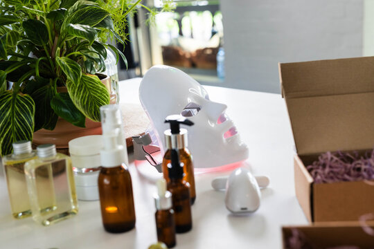 White LED facial mask glowing with pink light on tabletop near window, surrounded by amber bottles