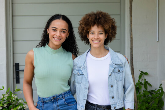 African American female friends standing on porch, smiling, in denim jacket and jeans near plants