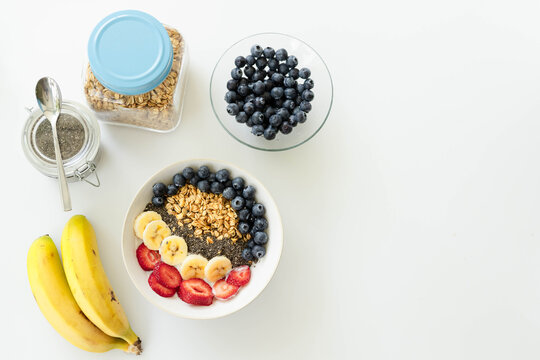 White ceramic bowl holding granola and fruit sitting left on white table near bananas, copy space