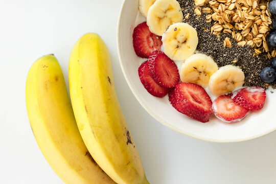 Yogurt bowl sitting on white table topped with granola, berries, chia, two bananas, copy space