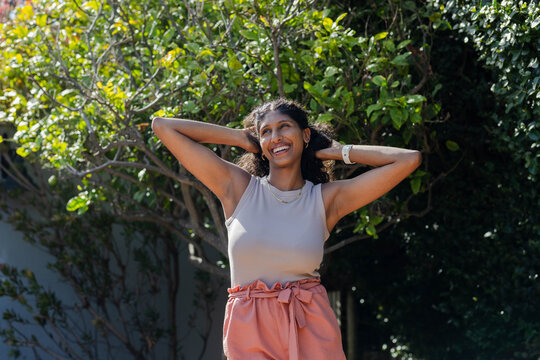 African American woman standing, arms raised, smiling in coral pants and gold necklace in backyard