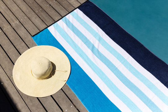 Striped beach towel lying flat on sunlit wooden deck, with straw hat near pool, showing shadows