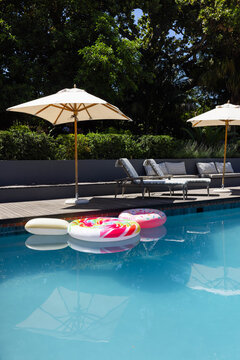 Three inflatable donut rings are floating in turquoise pool near wooden deck with beige umbrellas