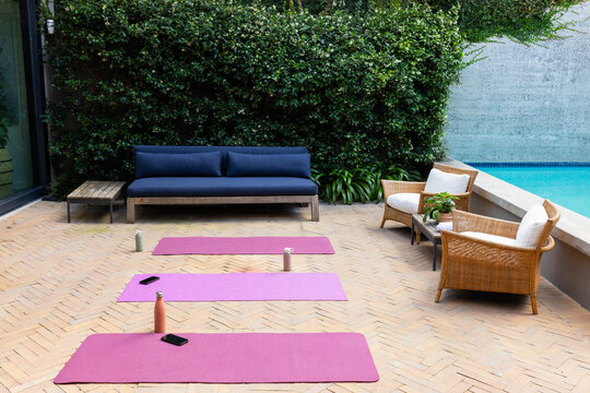 Three pink yoga mats are lying on herringbone patio by pool, with water bottles and phones