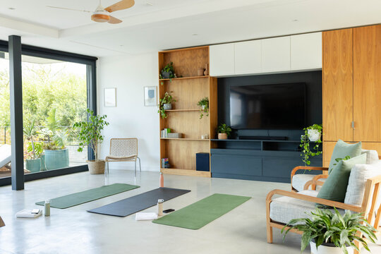 Three yoga mats are arranged on polished floor near sliding glass door, TV shelving, plants