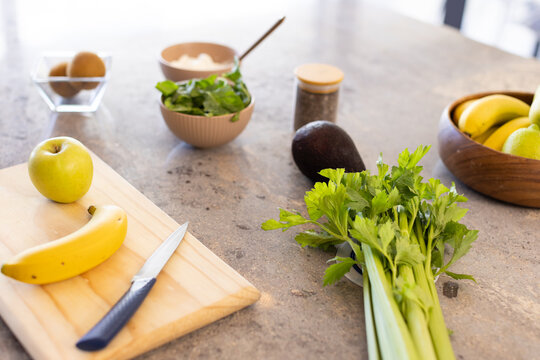 Bunch of celery lying on stone countertop, with cutting board holding banana, green apple, knife