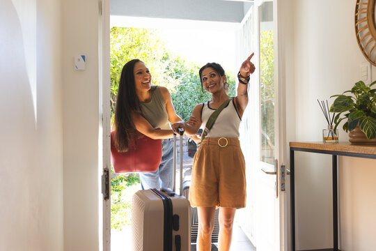 Medium wheeled suitcase rolling through open front door into sunlit entryway, red tote visible