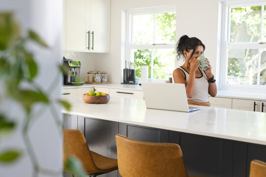 Woman standing at kitchen island wearing tank top holding mug near laptop, fruit bowl, copy space