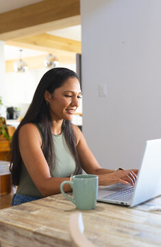 African American woman working on laptop at wooden kitchen table with green mug in olive top
