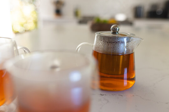 Glass teapot infusing amber tea, lid beading drops on marble counter near window, glass cup left