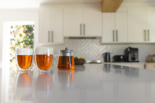 Two double-walled glass cups and glass teapot are reflecting amber tea on glossy white island