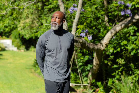 African man standing in sunlit garden near purple tree, wearing grey zip top and black pants