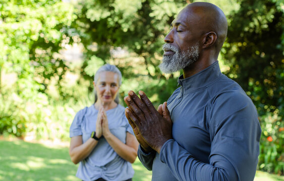 Diverse couple in activewear meditating in backyard senior man pressing hands woman with smartwatch