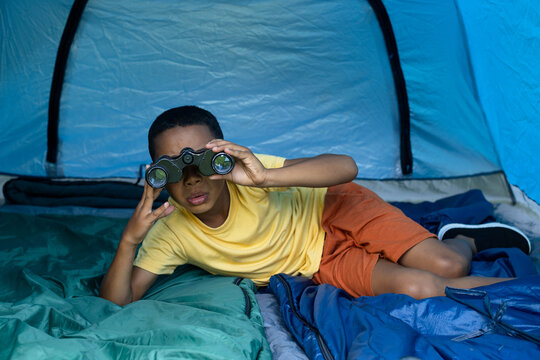 African teen lying on sleeping bags inside blue tent wearing yellow tee, holding binoculars