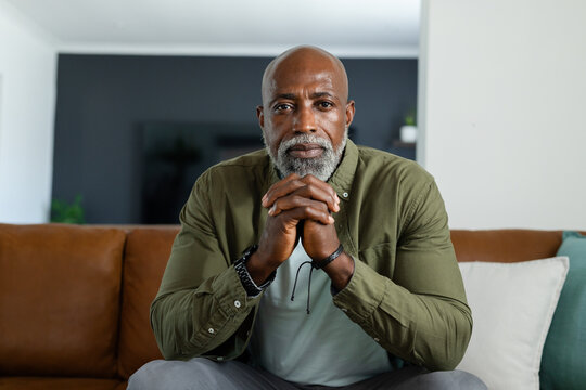 Mature African man sitting on leather sofa in living room, leaning forward clasping hands gazing