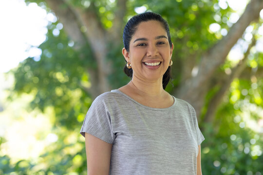Woman smiling and standing in park by tree, wearing light gray top and gold hoop earrings
