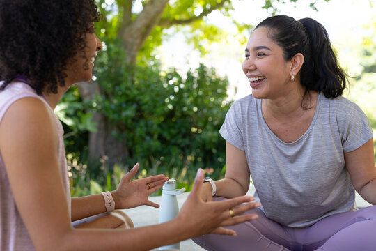 Diverse women friends laughing and talking sitting in park wearing activewear with water bottle