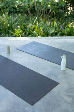 Dark gray yoga mat is lying diagonally on concrete patio with white bottle and green border
