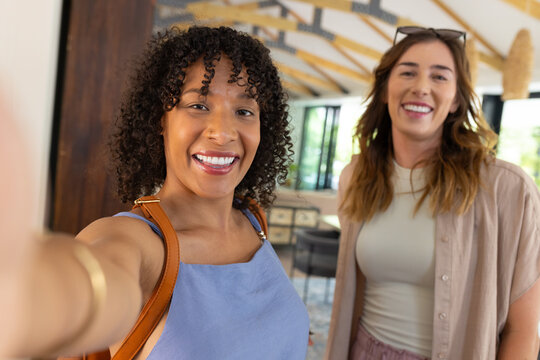 Diverse female friends posing holding smartphone for wrist selfie in bright lounge with beams