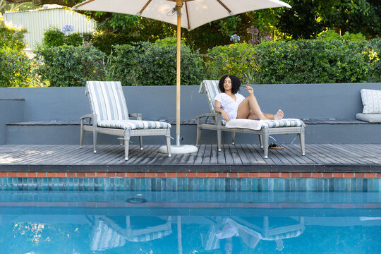 African American woman lounging on right chaise under umbrella by blue pool, sandals, copy space