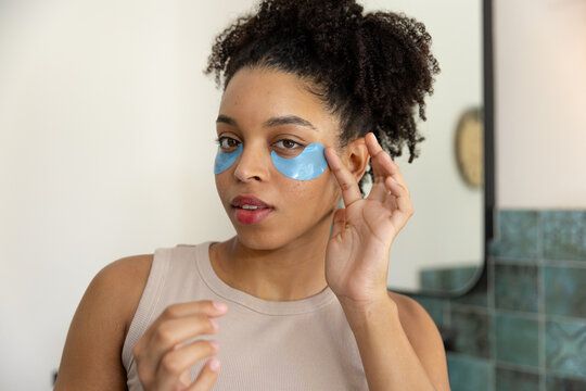 Adult African woman wearing beige tank applying blue hydrogel under-eye patches at bathroom mirror