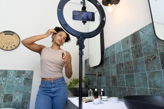 African American woman standing behind ring light applying drops from dropper at bathroom sink