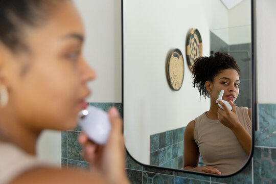African American woman in 20s using facial trimmer while looking into bathroom mirror, copy space