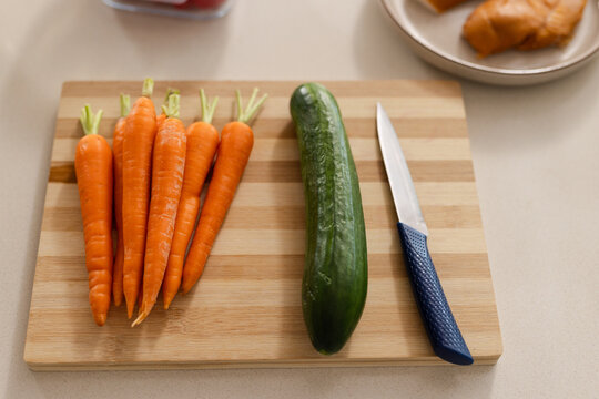 Wooden cutting board is holding seven carrots, cucumber and knife on light counter, pastry blurred