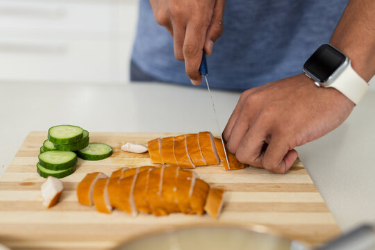 Blue-handled chef's knife is slicing cooked protein on wooden board on counter near cucumber rounds