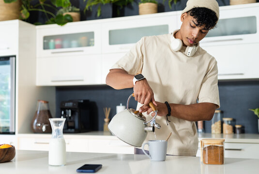 Man in beige shirt, beanie pouring kettle water into mug at kitchen island with smartphone