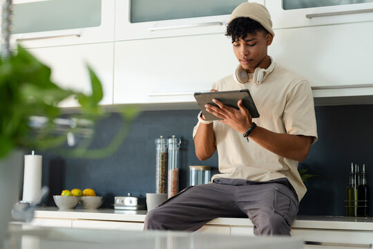 African American male early 20s sitting on kitchen counter using tablet wearing beanie, headphones